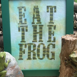 May include: A framed cross-stitch with the text "Eat the Frog" in green and yellow thread. Two green frog figurines are on either side of the frame.
