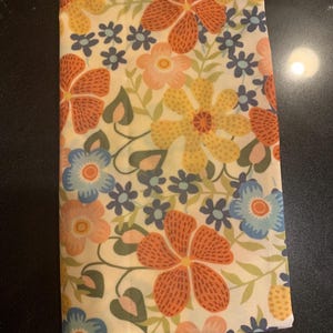 Op de afbeelding: Een witte stof met een bloemenpatroon in tinten oranje, geel, blauw en roze. De stof heeft een subtiele glans en een delicate textuur.