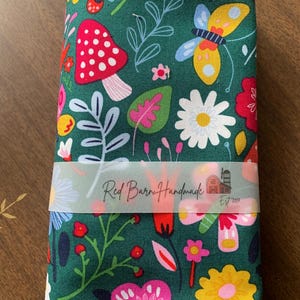 Op de afbeelding: Gevouwen stof met een donkergroene achtergrond, met een kleurrijk bloemen- en insectenpatroon. Het ontwerp bevat paddenstoelen, vlinders en diverse bloemen in tinten rood, roze, geel en wit. Een transparante band met de tekst "Red Barn Handmade Est 2017" wikkelt zich om de stof.