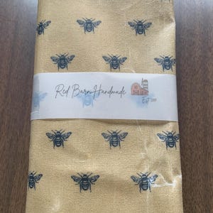 May include: A folded fabric with a repeating pattern of dark blue bees on a tan background. A white band across the middle reads "Red Barn Handmade Est 2017" with a small barn illustration.