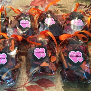 May include: A collection of cellophane bags filled with dried potpourri, secured with orange ribbons. Each bag features a sticker with "Joyce's Candles" and a pink heart. The potpourri contains dried flowers and spices, creating a fragrant mix.