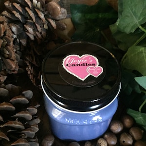 Op de afbeelding: Een blauwe glazen kaars pot met een zwarte deksel en een roze hartvormig label met de tekst "Joyce's Candles" met een kleiner hart met de tekst "Hand Poured with Love". De pot is omringd door dennenappels en eikels.