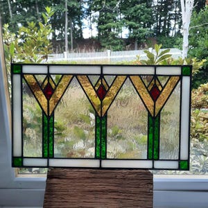 May include: Stained glass window panel featuring a geometric design with gold, red, green, and white glass. The panel has a rectangular shape and is displayed in front of a window, showcasing a garden view.