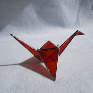 Op de afbeelding: Een glas-in-lood ornament in de vorm van een origami kraanvogel, gemaakt van doorschijnende rode glaspanelen die bij elkaar worden gehouden door dunne zilverkleurige metalen soldeerlijnen, met uitgestrekte vleugels en een puntige kop en staart.