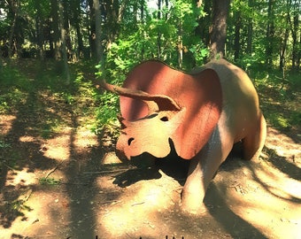 May include: A rusty brown metal sculpture of a triceratops dinosaur in a forest setting. The sculpture is partially obscured by trees and shadows.
