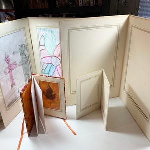May include: A set of five beige picture frames with a decorative border. The frames are arranged in a semi-circle on a white surface. The frames are empty except for one that contains a child's drawing of a flower.