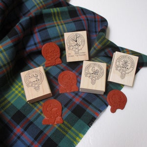 Pode incluir: Cinco carimbos de madeira com diferentes desenhos e texto, incluindo "An Cuanan", "Committ Thy Work", "Aonaidh Ri Cheile" e "Clan Grant", em um tecido xadrez verde, azul e amarelo.