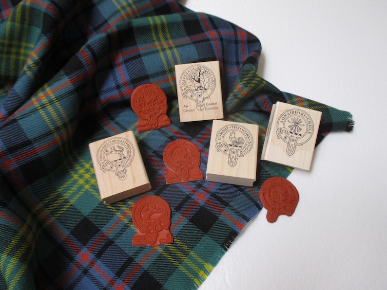 Pode incluir: Cinco carimbos de madeira com diferentes desenhos e texto, incluindo "An Cuanan", "Committ Thy Work", "Aonaidh Ri Cheile" e "Clan Grant", em um tecido xadrez verde, azul e amarelo.