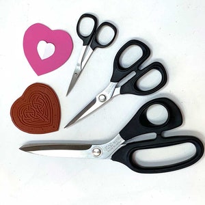 May include: An assortment of craft supplies including three pairs of scissors with black handles and silver blades, a pink heart with a white heart cutout, and a brown heart-shaped stamp with a Celtic knot design.