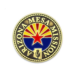 May include: A gold-colored metal coin with the words "Arizona Mesa Mission" around the edge. The center of the coin features a red star on a blue background with a yellow and red sunburst design behind it. The bottom of the coin features a silhouette of the state of Arizona with a cactus.