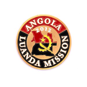 May include: A gold-colored round pin with the words "Angola Luanda Mission 2013" around the edge. The center of the pin features a map of Angola with the Angolan flag colors of red, black, and yellow.