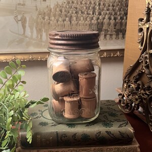 May include: A clear glass Mason jar filled with vintage wooden spools. The jar has a rusty metal lid and is sitting on top of two antique books with green covers. A framed black and white photograph is in the background.