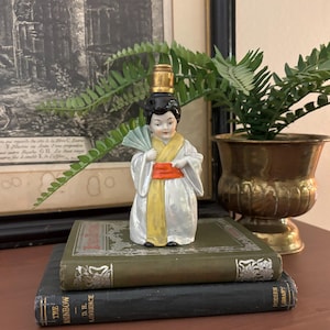 May include: A small porcelain figurine of a woman in a traditional Japanese kimono, holding a fan. The kimono is white with yellow and orange accents. The figurine has black hair and is topped with a gold-colored stopper. It is displayed on a stack of vintage books.