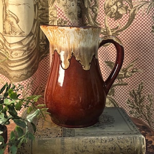 May include: A ceramic pitcher with a rich, glossy brown glaze. The top of the pitcher features a dripped, creamy white and tan glaze. It has a curved handle and a spout for pouring. The pitcher is likely used for serving beverages.