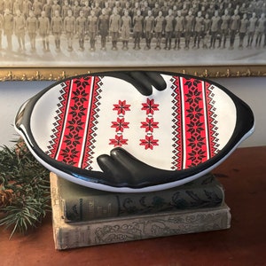 May include: A white ceramic serving dish with black hand-shaped handles. The dish is decorated with a red and black geometric pattern.