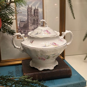 May include: A white porcelain sugar bowl with a lid and floral design. The bowl has two handles and a scalloped edge. It is sitting on top of two books.