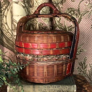 May include: A round, woven basket with a handle, featuring a red and brown color scheme. The basket has a lid and is sitting on top of a stack of books. The handle is a dark red color.