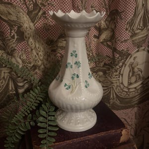 May include: White ceramic candlestick holder with a scalloped top and a flared base. The holder is decorated with green shamrock designs. The candlestick holder is displayed on top of a stack of antique books.