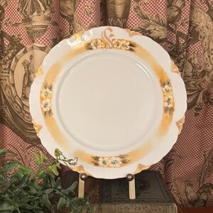 May include: A white porcelain plate with a scalloped edge, featuring a decorative border of orange and gold. The plate is adorned with floral accents and is displayed on a gold-toned stand. The plate's design is reminiscent of vintage tableware.