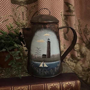 May include: A vintage copper-toned metal coffee pot with a painted coastal scene. The artwork depicts a lighthouse, sailboat, and clouds against a blue and white background. The pot has a handle, spout, and a lid with a small loop.