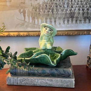 May include: A green ceramic figurine of a person wearing a hat sits on a green ceramic leaf-shaped dish. The figurine and dish are on top of a stack of three vintage books.