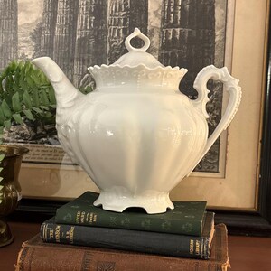 May include: A white porcelain teapot with a decorative lid and handle. The teapot is placed on a stack of vintage books with green and brown covers. The background features a framed print of classical architecture.