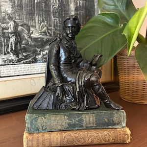 May include: A bronze-colored statue of a seated man reading, atop a stack of two antique books. The books are green and tan with gold lettering. The background includes a framed black and white print and a potted plant.