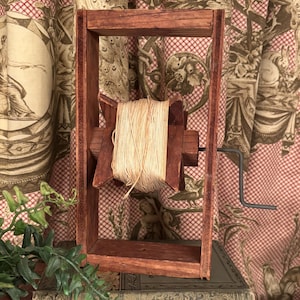 May include: A vintage wooden yarn swift with a spool of natural-colored yarn. The swift is a dark reddish-brown color and has a hand crank. The yarn is tightly wound around the spool. The swift sits on top of a stack of old books.
