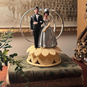 May include: A vintage wedding cake topper featuring a bride and groom figurine. The groom wears a black tuxedo, and the bride has a silver dress. The figurines stand in front of a silver ring. The topper sits on a cream-colored, flower-shaped base.