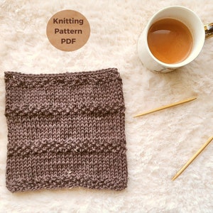 May include: A brown knitted fabric swatch with a chevron pattern. The fabric is on a white fluffy surface with a cup of tea and knitting needles.