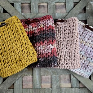 May include: Four handmade crochet pouches in mustard yellow, burgundy, light pink, and lavender. Each pouch features a wooden button closure. The pouches are arranged on a rustic wooden background, showcasing the texture and colors of the yarn.