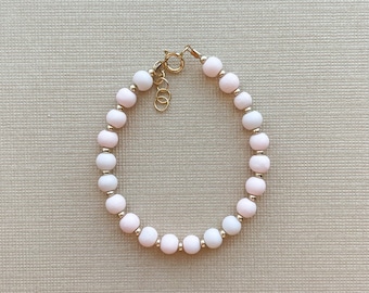 Puede incluir: Un delicado brazalete hecho de cuentas blancas y rosadas con un cierre dorado.