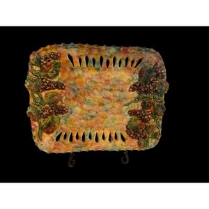 May include: Rectangular decorative tray with a textured surface and a colorful, mottled design. The tray features grape clusters and vines in relief around the edges, with a series of cut-out slits. The tray is supported by a dark metal stand.