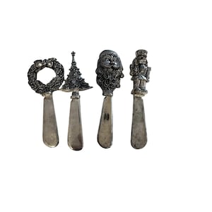 May include: A set of four silver-coloured Christmas spreaders. Each spreader has a different festive design: a wreath, a Christmas tree, Santa Claus, and a nutcracker. The spreaders have a classic, vintage appearance.