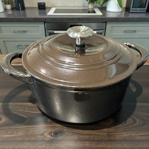 Op de afbeelding: Een donkerbruine gietijzeren braadpan met een bijpassende deksel en een zilveren knop. De pot heeft twee grote handvatten en staat op een houten oppervlak. Het kookgerei is ontworpen om te koken.
