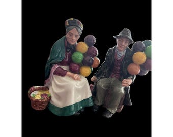 May include: A ceramic figurine set depicting two figures, a woman and a man, each holding a cluster of colorful balloons. The woman is seated, wearing a headscarf and apron, with a basket of flowers nearby. The man is seated, wearing a hat and coat.