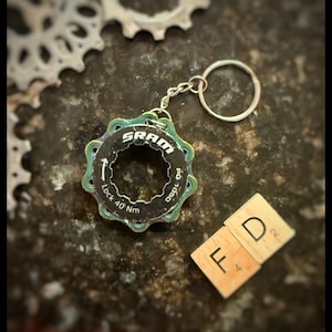 May include: A bicycle gear-themed keychain with a silver keyring. The keychain features a black and iridescent green SRAM gear component with the text "SRAM" and "Lock 40 Nm". Two wooden tiles spell "FD".