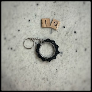 May include: A black metal keychain shaped like a bicycle chain with a silver ring. The keychain is on a white surface with two wooden letter tiles that spell "IG".
