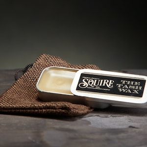 May include: A silver tin with a black label that reads "Squire The Gentleman's Choice Original Product The Tash Wax". The tin is open and contains a small amount of yellow wax.