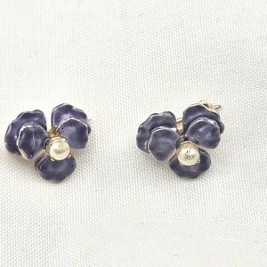 May include: A pair of flower-shaped earrings. Each earring features three dark purple petals surrounding a cream-colored pearl. The earrings have a gold-colored metal base and are set against a white background.