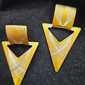 May include: A pair of gold triangle earrings with a white geometric design. The earrings have a textured surface and a shiny finish.