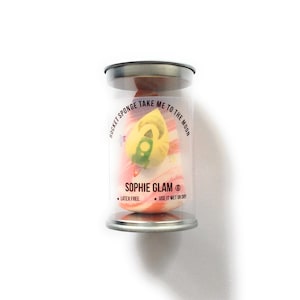 May include: A pink and yellow makeup sponge in a clear plastic container with a silver lid. The sponge is shaped like a rocket and has the text "ROCKET SPONGE TAKE ME TO THE MOON" and "SOPHIE GLAM®" printed on it. The container also has the text "LATEX FREE" and "USE IT WET OR DRY" printed on it.