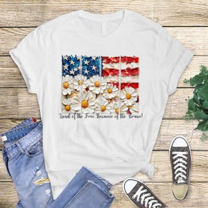 May include: White t-shirt featuring a patriotic design with an American flag motif. The flag design incorporates white daisies and the text "Land of the Free, Because of the Brave!"