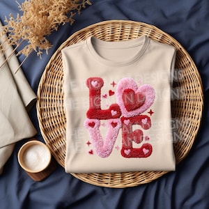 May include: A cream-colored sleeveless shirt with the word "LOVE" in a textured, plush design. The letters are red and pink, with heart and star accents. The shirt is folded on a woven basket.