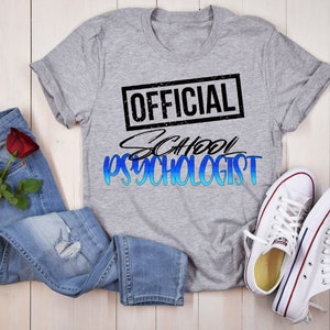 May include: A grey t-shirt with the text "OFFICIAL SCHOOL PSYCHOLOGIST" in black and blue lettering. The text is in a distressed font style.