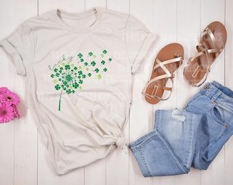 Puede incluir: Una camiseta beige claro con un diseño de diente de león de trébol verde y el texto "Lucky" impreso en la parte delantera. La camiseta está colocada sobre una superficie de madera blanca con un par de sandalias doradas y unos vaqueros azules.