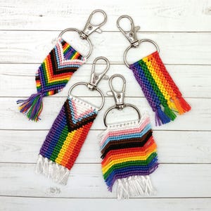 May include: Four handmade keychains with silver-colored clasps. Each keychain features a woven design in vibrant rainbow colors, with a different pattern on each. The keychains have a fringe of matching colored threads at the bottom, and are displayed on a light wood surface.