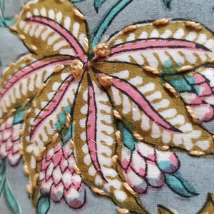 May include: Close-up of a blue fabric with a floral pattern. The fabric features a large, pink and white flower with gold embroidery. The flower has a detailed design with intricate stitching.