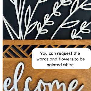 May include: Wooden welcome sign with white floral accents and the word "welcome" in white script. The sign has a dark background and a decorative wooden border. Text on the sign reads: "You can request the words and flowers to be painted white."