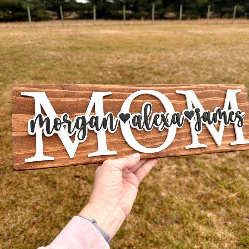 May include: A wooden sign with the word "MOM" in large white letters. The letters are raised and have a rustic look. The sign also has the names "Morgan", "Alexa", and "James" written in black cursive with a small black heart between each name.